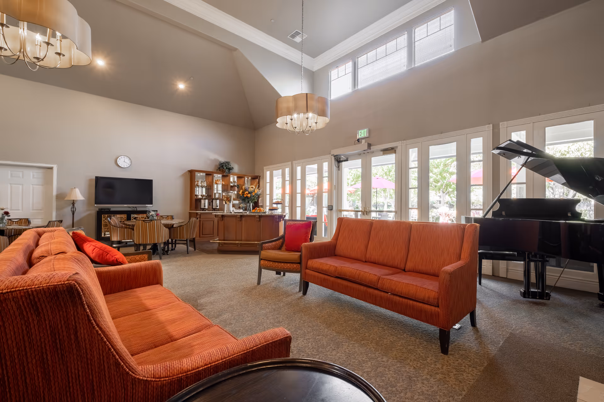 Spacious senior living common room with red sofas, a grand piano, dining area and large windows letting in natural light.