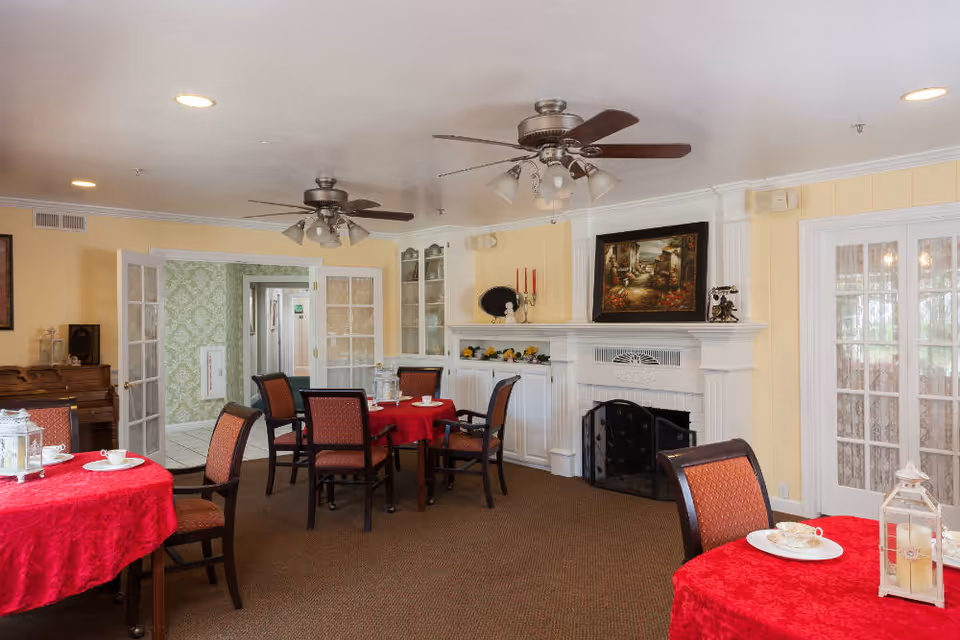 Dining room with round tables covered in red tablecloths, upholstered chairs, a white fireplace mantel, ceiling fans, and French doors.