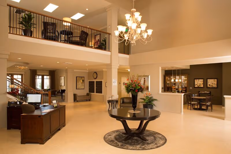 Spacious two-story lobby with a central round table and floral arrangement, chandelier, reception desk, seating areas, and an upper balcony.