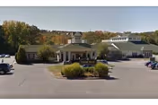 Exterior view of Cheshire House senior living facility with a large parking lot in front, surrounded by trees with autumn foliage.