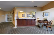 Reception area with a wooden front desk and a glass partition. To the right, there is a wooden table with two chairs and a lamp. The walls are painted beige with framed artwork and a crucifix on the left wall. The floor is carpeted with a patterned design.