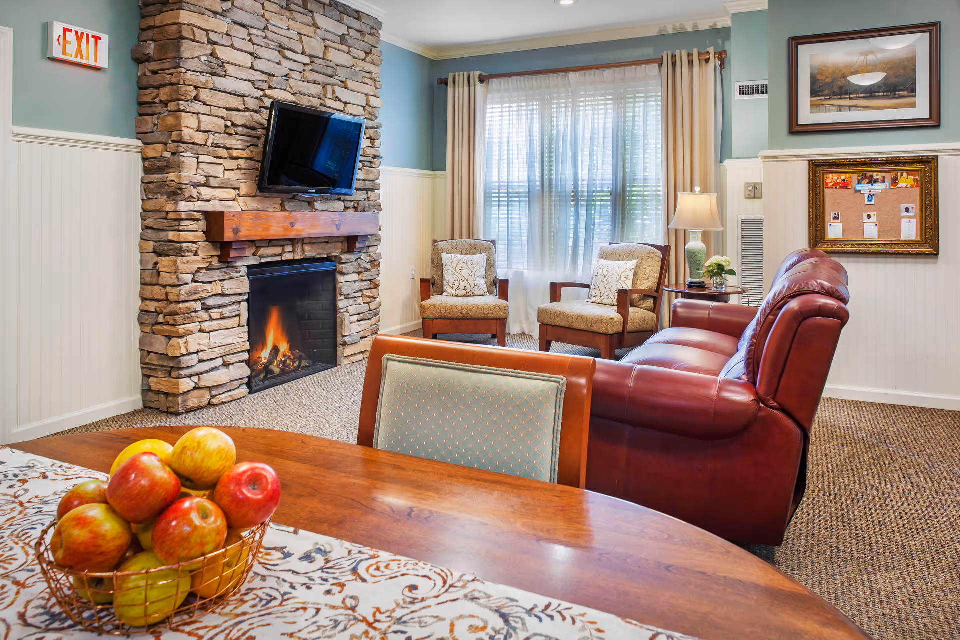 Cozy lounge area with a stone fireplace, wall-mounted TV, leather sofa, upholstered chairs, and a dining table with a bowl of apples.