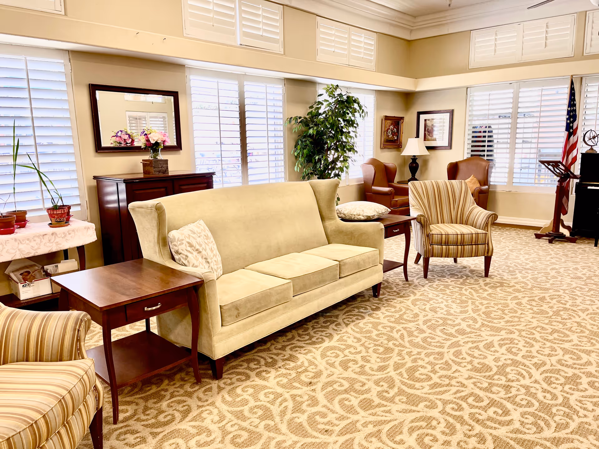 A bright and spacious living room in an assisted living facility featuring a beige sofa with patterned pillows, two striped armchairs, two brown leather armchairs, a wooden side table, a lamp, framed artwork on the walls, a large green potted plant, and windows with white plantation shutters allowing natural light to fill the room. The floor is covered with a patterned carpet.