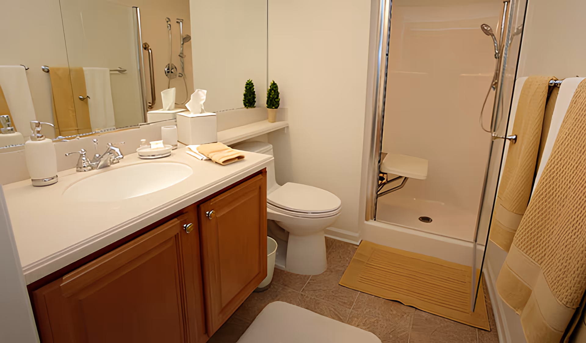 A clean and well-organized bathroom featuring a sink with a countertop, a large mirror, a toilet, and a walk-in shower with a foldable seat. The bathroom has beige and white tones with matching towels hanging on a rack and a small potted plant on a shelf above the toilet.