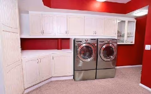 Laundry room with red walls, beige carpet, white cabinets, and a front-loading washer and dryer set in stainless steel finish.