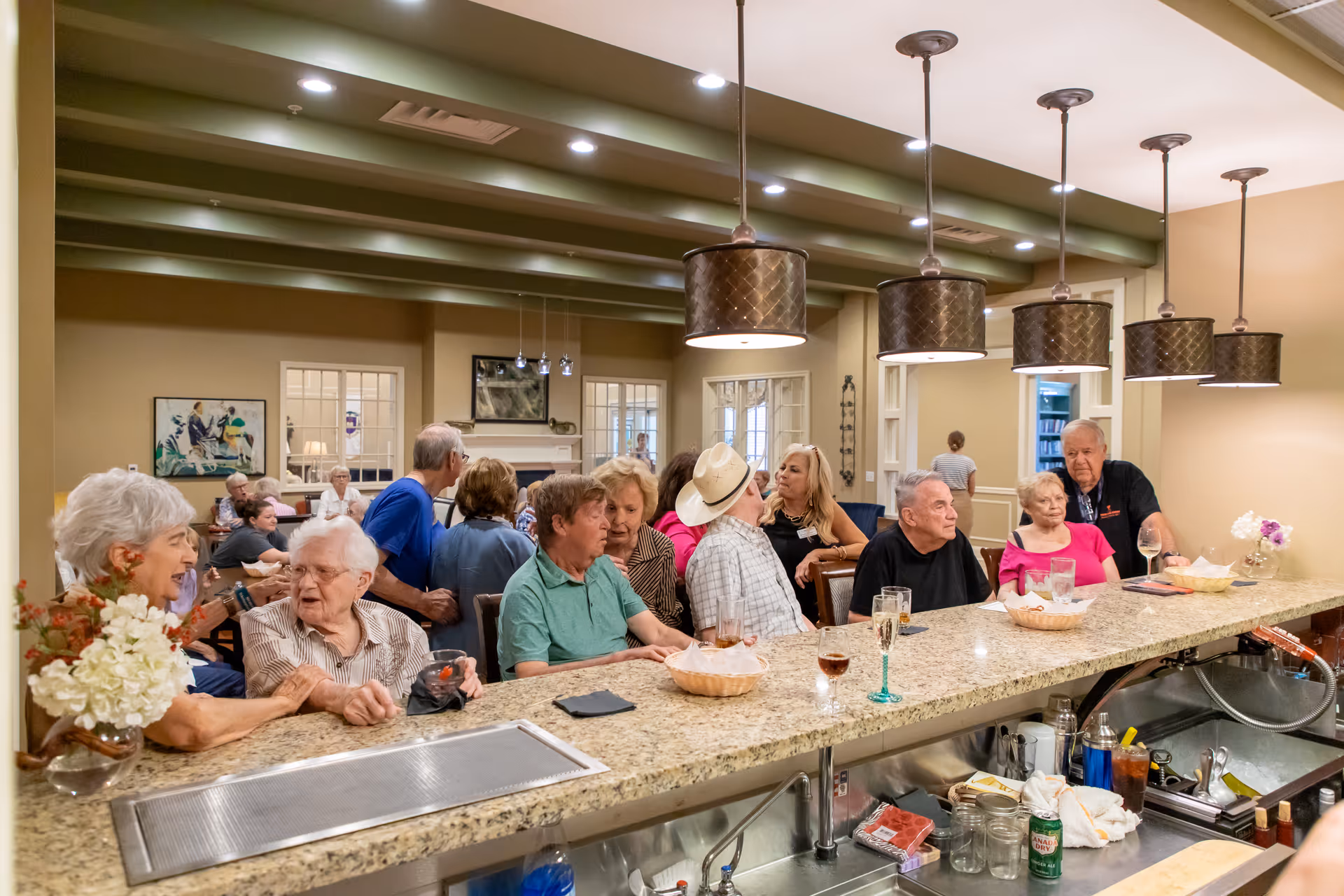 Several elderly residents sit and socialize along a granite countertop bar in a bright communal room.