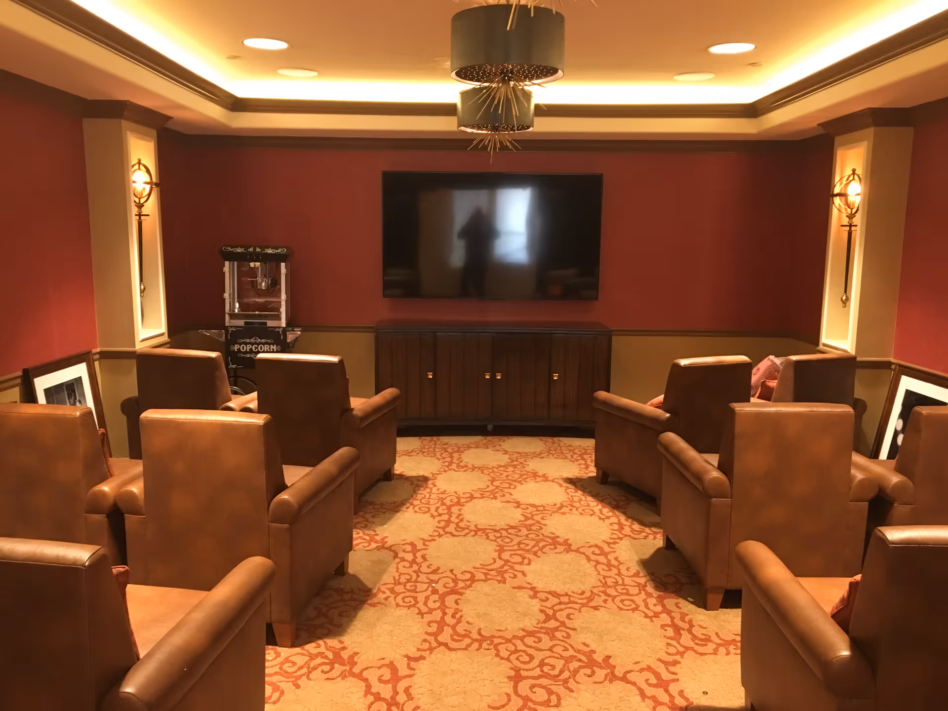 A cozy media room with eight brown leather armchairs arranged in two rows facing a large flat-screen TV mounted on a red and beige wall. There is a popcorn machine in the corner and decorative lighting fixtures on the walls. The room has a patterned carpet and a ceiling with recessed lighting.