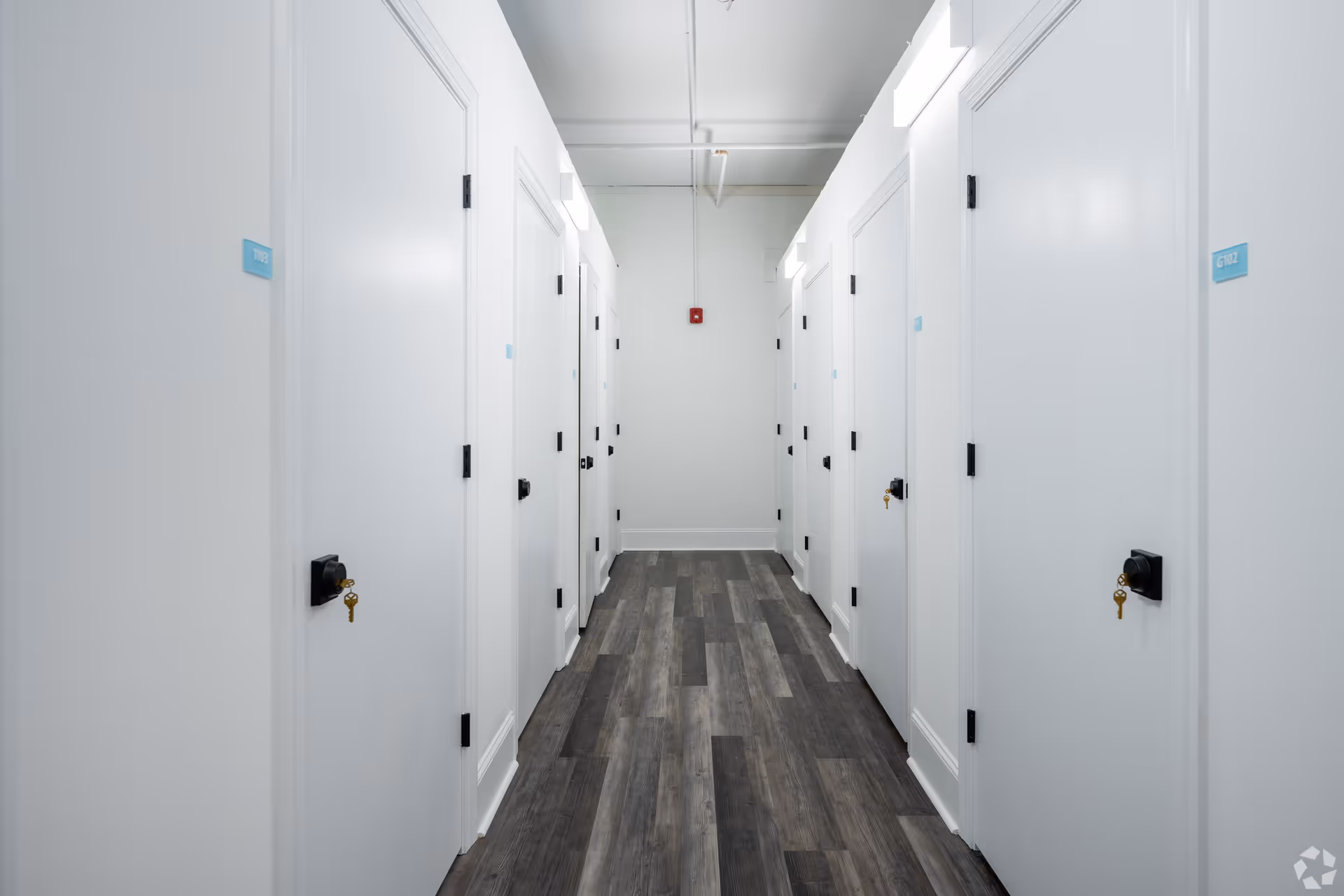 A clean, well-lit hallway with white walls and multiple white doors on both sides, each door having a black lock with a key hanging from it. The floor is covered with dark wood-patterned vinyl planks, and there are small blue room number signs on each door. The ceiling has exposed pipes and modern light fixtures.