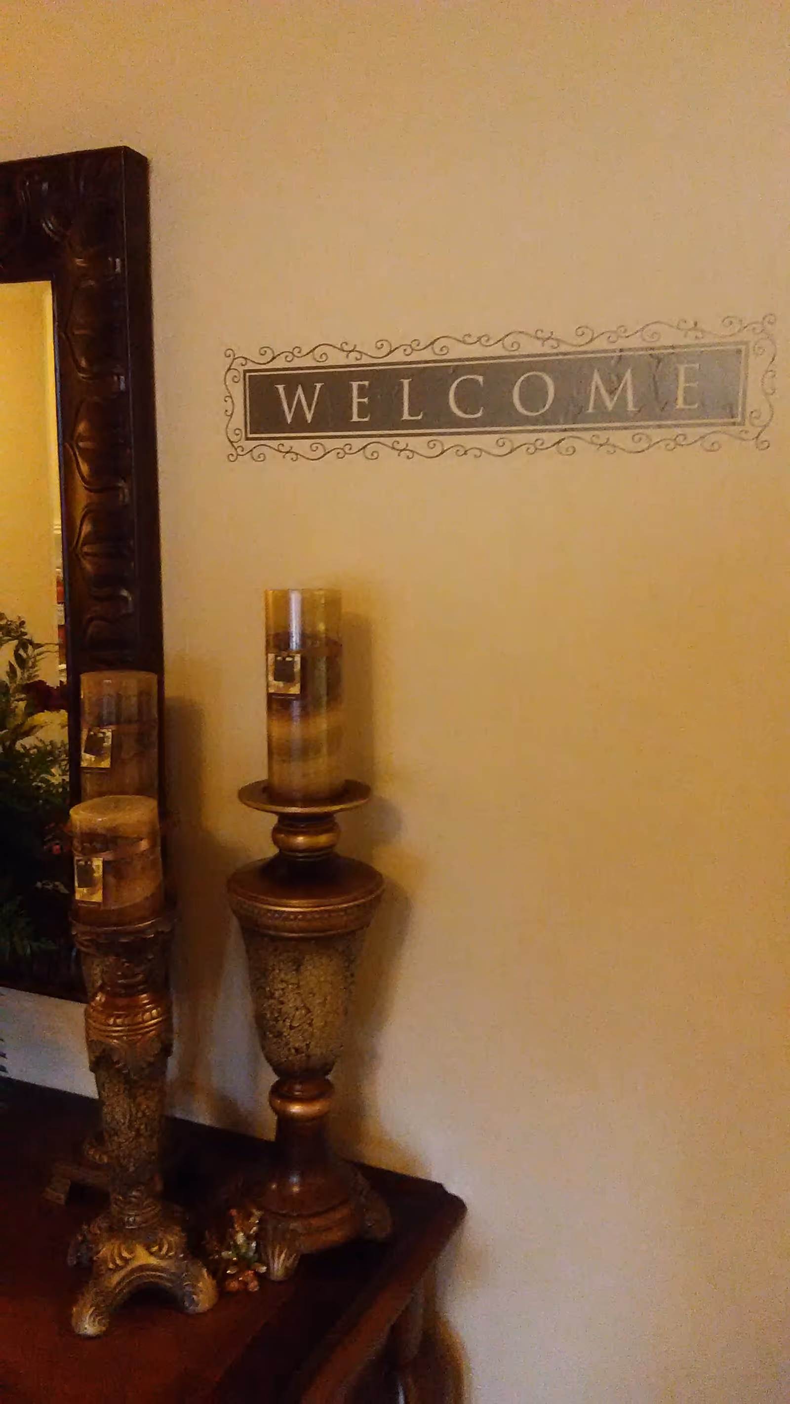 A decorative corner of a room featuring a large ornate mirror, two tall decorative candle holders with candles, and a wall decal that says 'WELCOME' in a framed design.