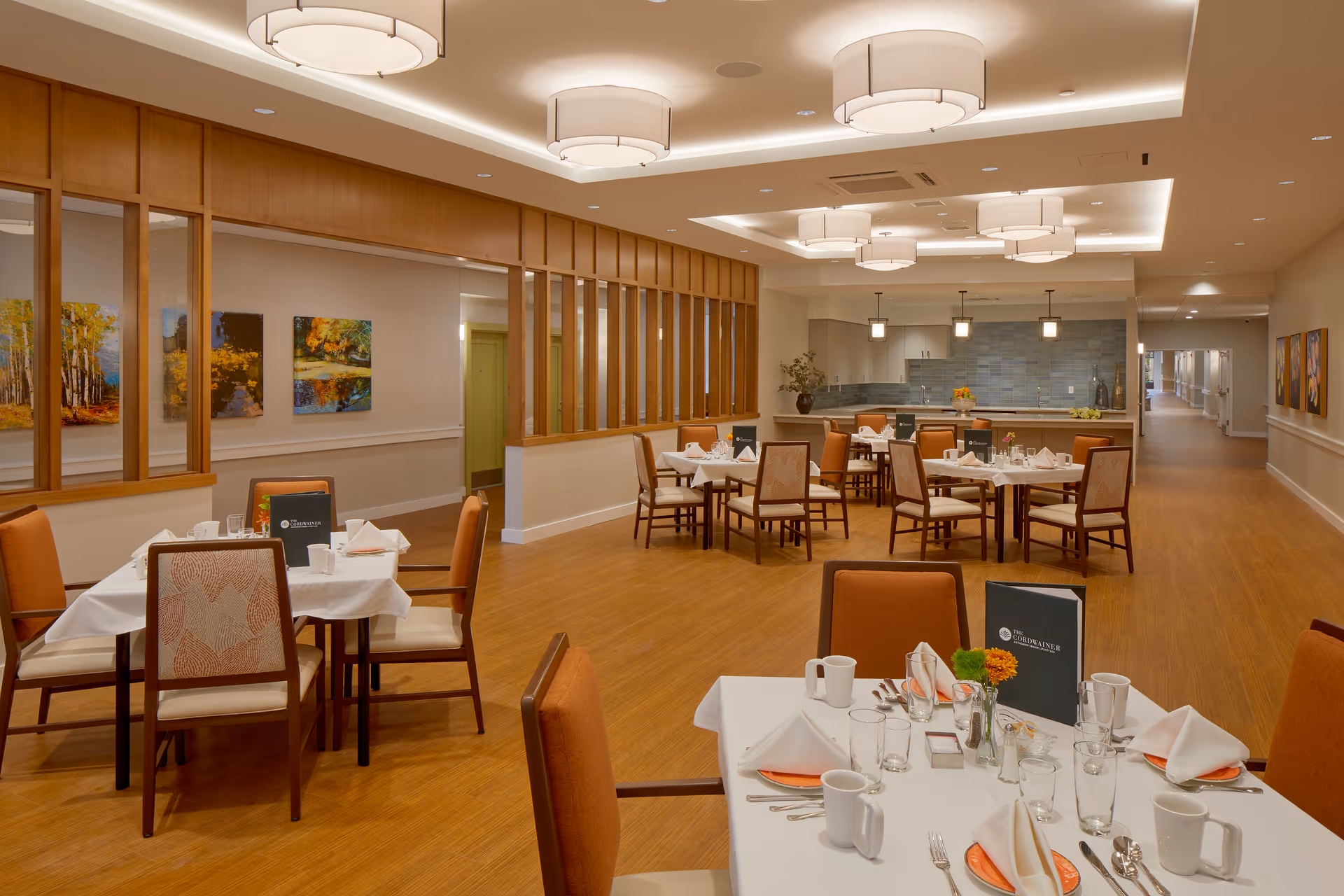 A well-lit dining room with multiple tables set with white tablecloths, napkins, plates, cups, and silverware. The chairs have orange cushions and patterned backs. The room features wooden flooring, modern ceiling lights, and a partial wooden divider with vertical slats. There are paintings on the walls and a kitchen area visible in the background.