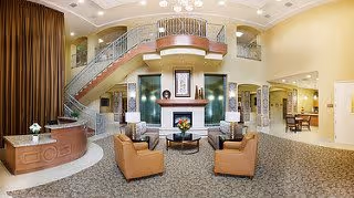 Spacious and well-lit senior living facility lobby with a seating area consisting of four tan armchairs around a coffee table in front of a fireplace. A curved staircase with metal railings leads to an upper level. To the left is a reception desk, and to the right, a hallway and dining area are visible. The walls are painted in warm tones, and the floor is carpeted with a patterned design.