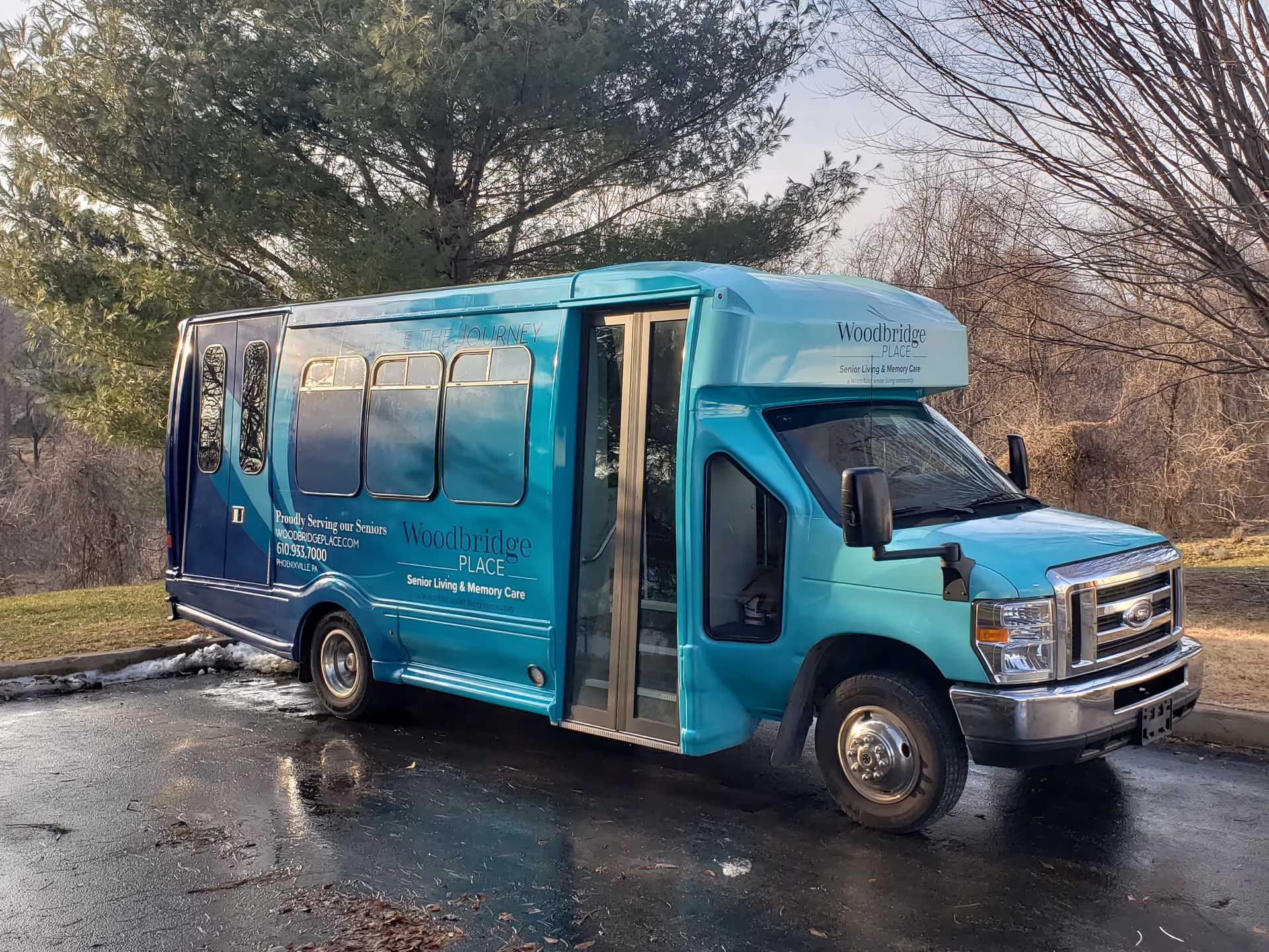 A teal and blue shuttle bus parked on a wet driveway near trees. The bus has signage for Woodbridge Place Senior Living & Memory Care, including contact information and the slogan 'Proudly Serving our Seniors.'
