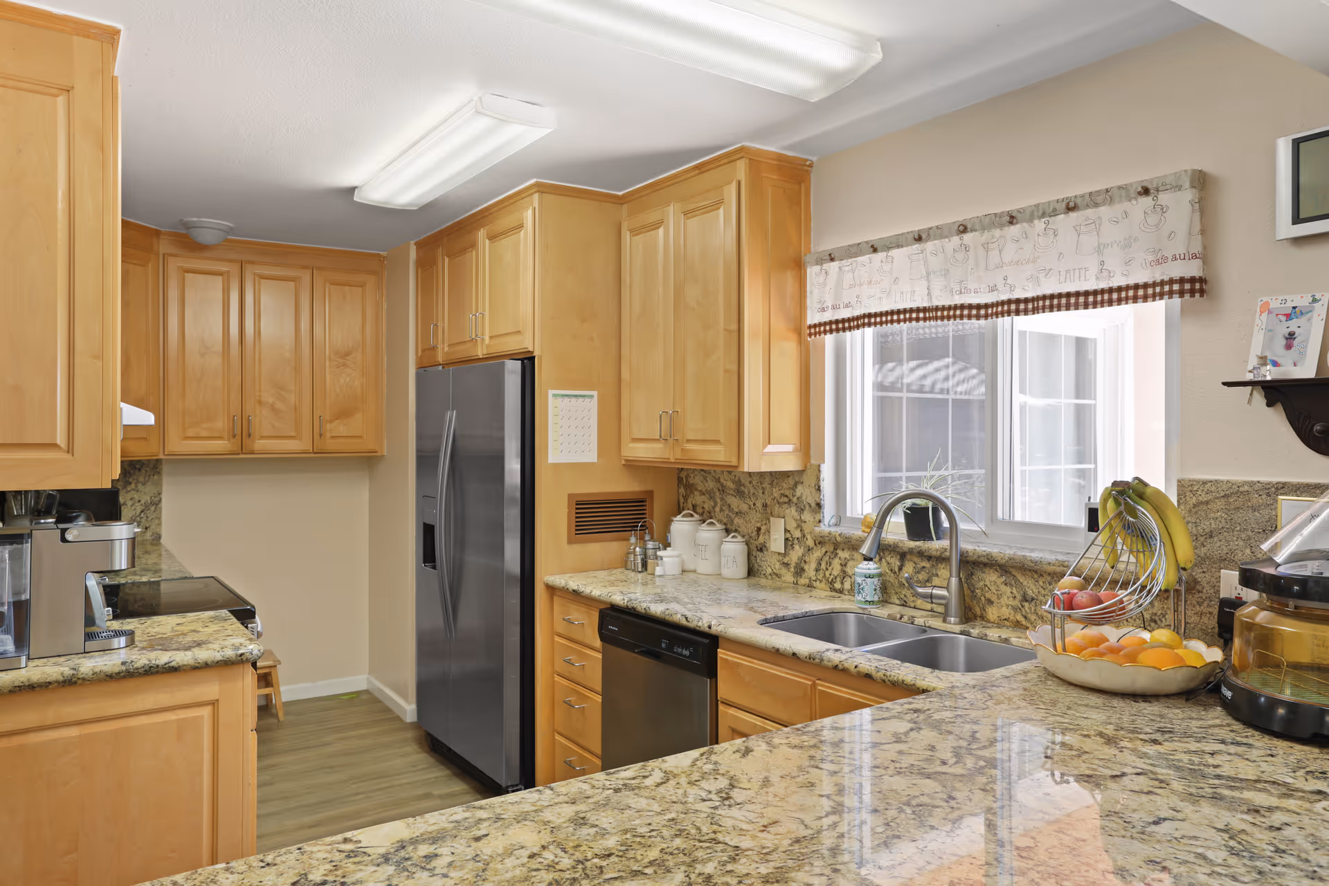 Bright kitchen with light wood cabinets, granite countertops, a stainless steel refrigerator, double sink under a window, and fruit on the counter.