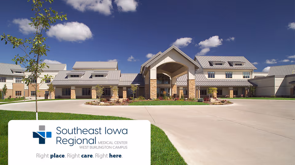 Front entrance of a large modern medical building with a covered porte-cochere, circular driveway, landscaping, and a bright blue sky.