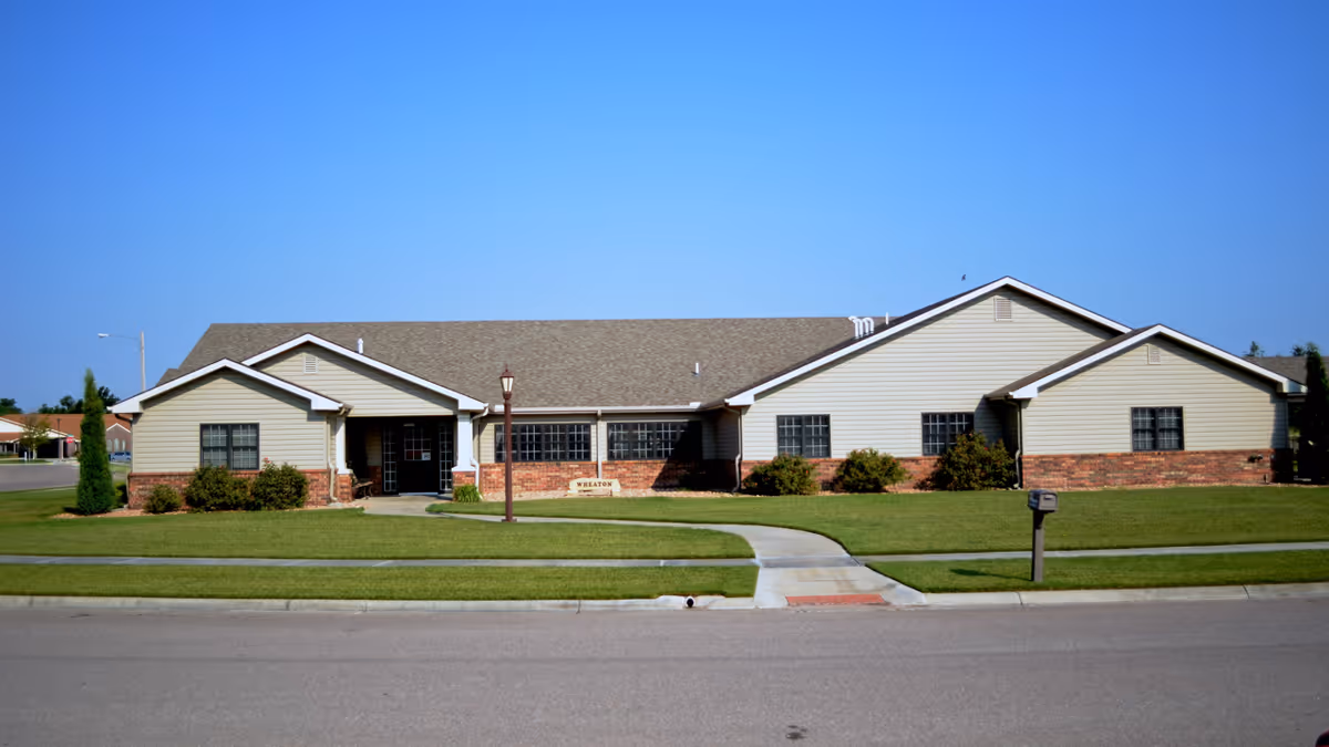 Single-story ranch-style building with beige siding and brick base, a front lawn, walkway and mailbox under a clear blue sky.