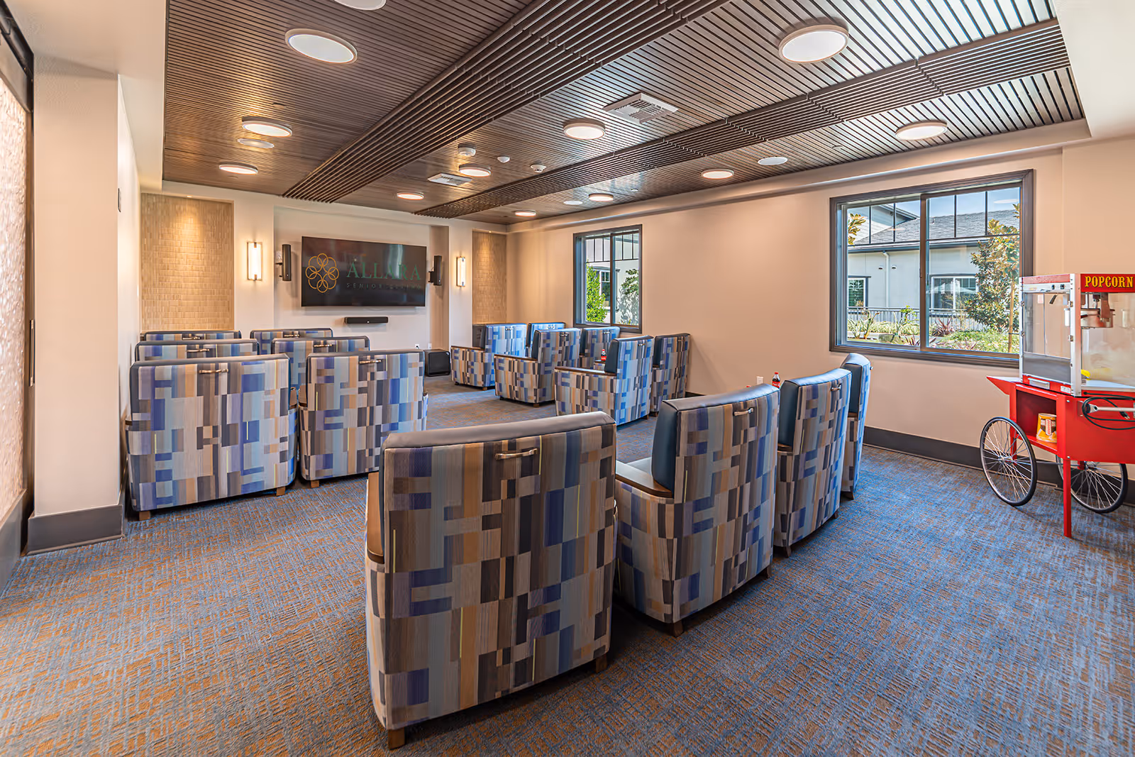 A cozy senior living common room with multiple patterned armchairs arranged in rows facing a large wall-mounted TV displaying the Allara Senior Living logo. The room features a modern wooden slatted ceiling with recessed lighting, large windows letting in natural light, and a red popcorn machine on the right side near the window.