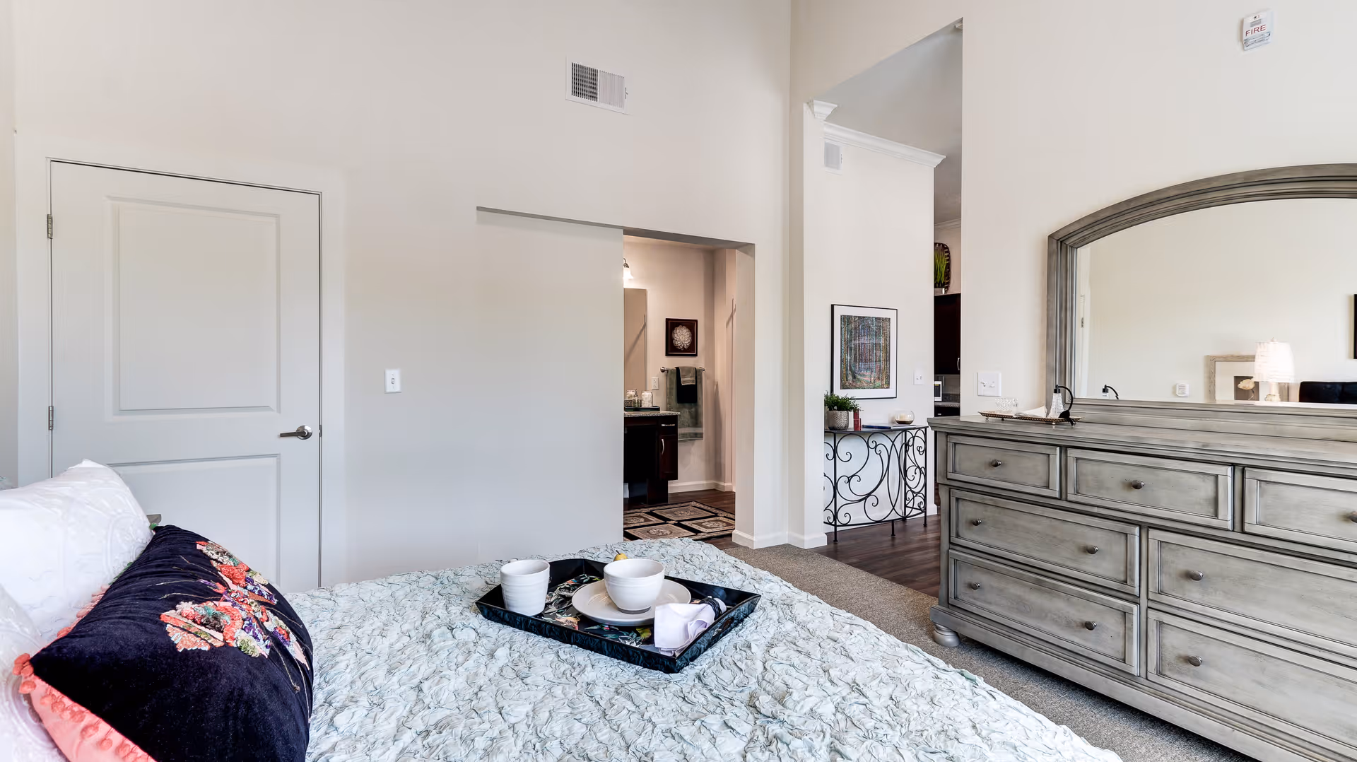 A bedroom in Ridgemere Senior Living featuring a bed with a floral pillow and a tray with cups and a bowl. The room has light-colored walls, a door, a dresser with a large mirror, and an open doorway leading to a bathroom and another room.