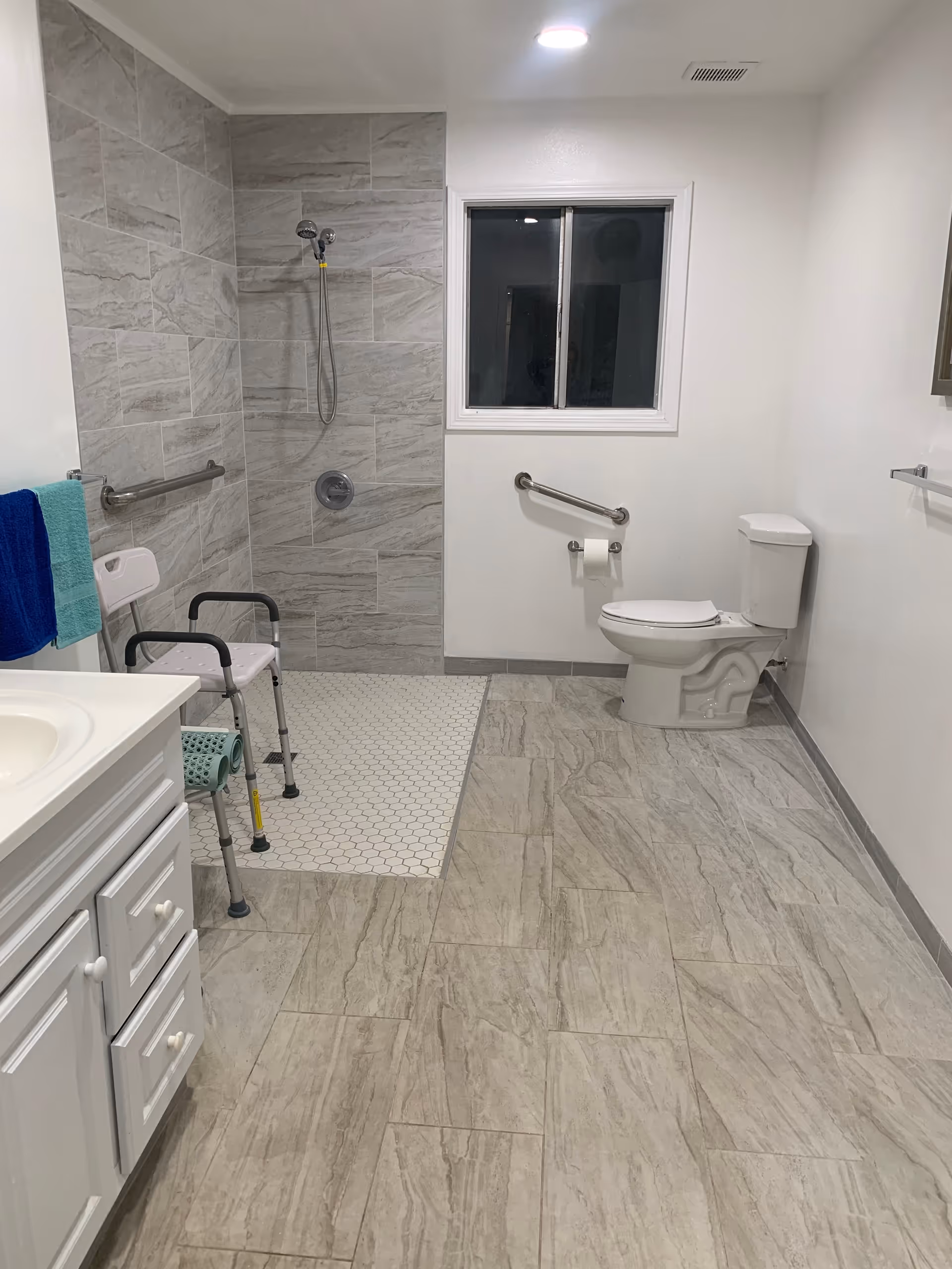 A spacious bathroom with light gray tiled floor and walls. It features a walk-in shower with a handheld showerhead, a white shower chair, grab bars on the walls, a toilet with a grab bar beside it, a white vanity with a sink, and a window above the toilet. Towels hang on a bar near the vanity.