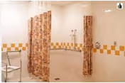 A bathroom with a shower area featuring a wall-mounted showerhead and two floral-patterned shower curtains. There is a metal chair with armrests positioned near the shower. The walls are tiled with a white and yellow checkerboard pattern along the middle.