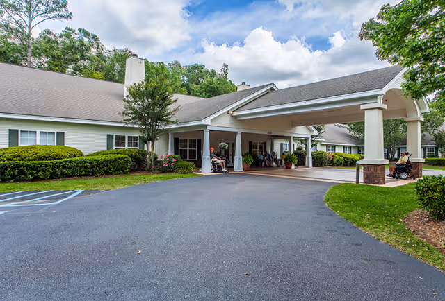 Exterior view of Oaks at Charleston senior living facility showing a single-story building with a covered entrance driveway. Several people are seated near the entrance, including one person in a wheelchair. The area is surrounded by greenery and trees under a partly cloudy sky.