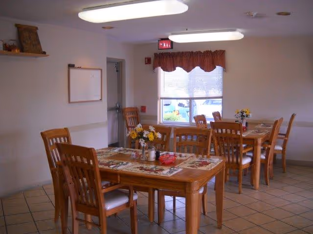 A dining room with wooden tables and chairs arranged neatly. Each table has placemats and small flower arrangements. There is a window with a red and white checkered valance, and fluorescent ceiling lights illuminate the room. A whiteboard and a small shelf with decorative items are mounted on the wall.