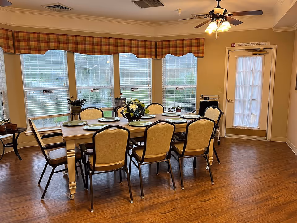 A dining room with a rectangular wooden table set with eight plates and a floral centerpiece. Eight cushioned chairs surround the table. The room has large windows with white blinds and plaid valances, a wooden floor, a ceiling fan with lights, and a door with a sheer curtain.