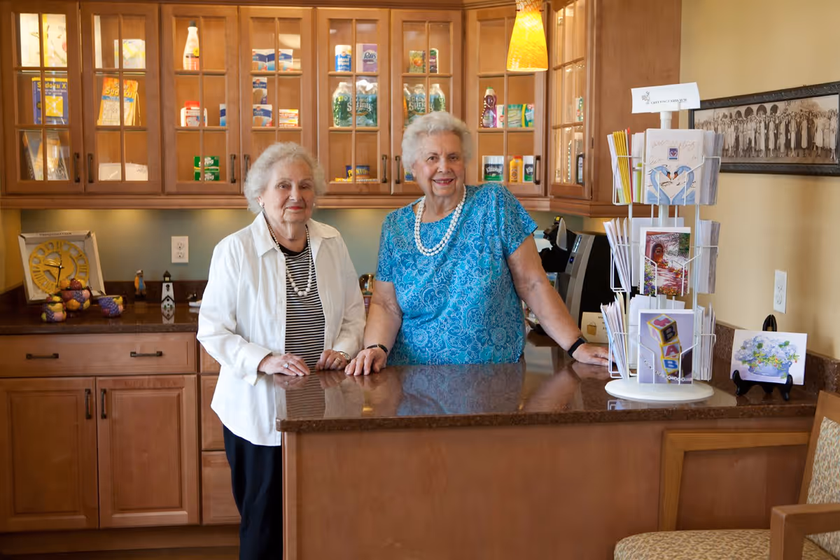 Two elderly women standing behind a kitchen counter in a well-lit room with wooden cabinets filled with various items. One woman is wearing a white jacket over a striped shirt and the other is wearing a blue patterned blouse with a pearl necklace. There is a rotating card display on the counter and a framed black and white photo on the wall.