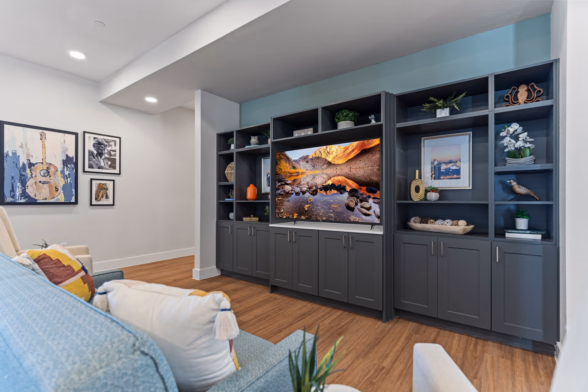 A cozy living room area featuring a large built-in dark gray shelving unit with a flat-screen TV displaying a scenic mountain and lake view. The shelves are decorated with plants, framed pictures, and decorative items. In the foreground, there is a blue couch with colorful pillows and a beige armchair. The room has wooden flooring and white walls with framed artwork hanging on one side.