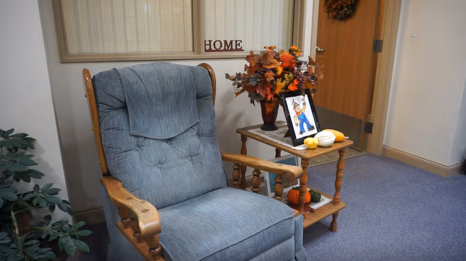 Blue upholstered recliner beside a wooden side table topped with autumn flowers, small pumpkins and a framed picture in an indoor common area.