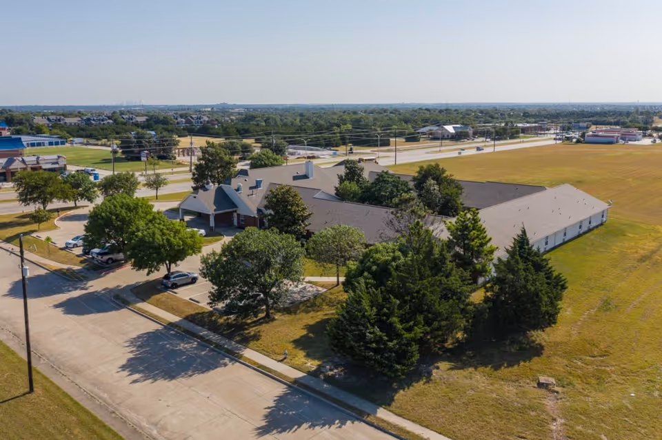 Aerial view of Cedar Hill Senior Living facility showing a large single-story building surrounded by trees and parking spaces, with open grassy areas and roads nearby under a clear sky.