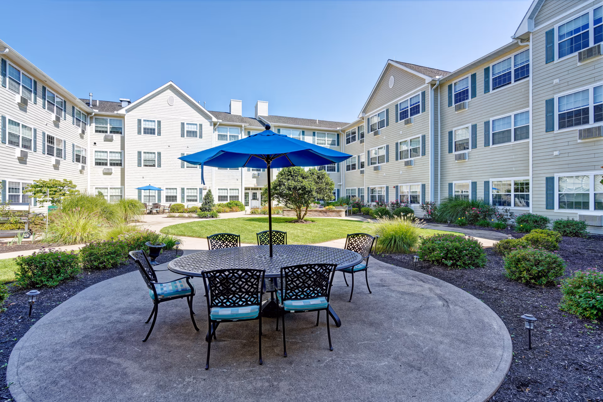 Sunlit courtyard patio with a round table, blue umbrella and chairs surrounded by landscaping and three‑story residence buildings.
