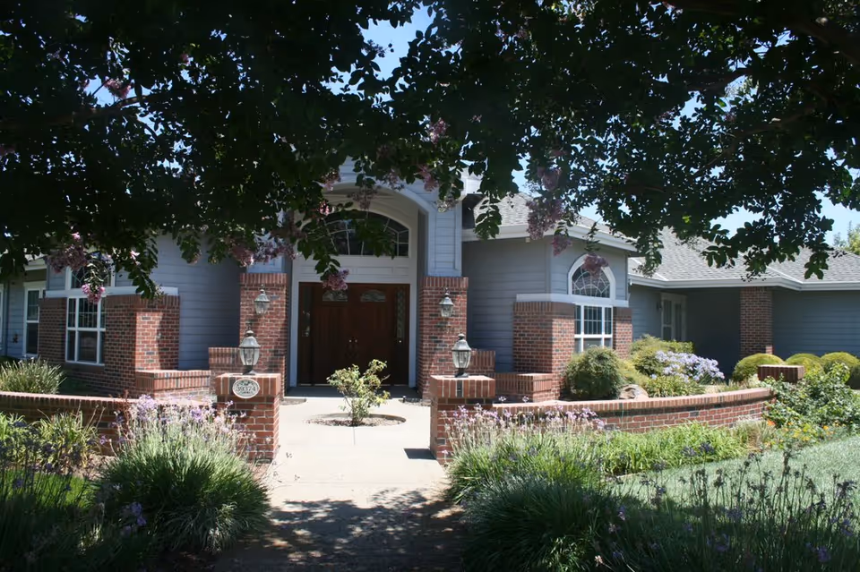 Front exterior view of a single-story building with gray siding and red brick accents, featuring a central entrance with double wooden doors and arched windows above. The entrance is framed by brick pillars with lantern-style lights. The foreground includes a garden with green shrubs, flowering plants, and a tree with hanging purple flowers providing shade.