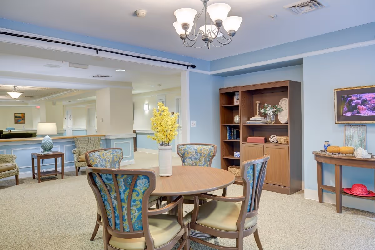 A cozy interior common area with a round wooden table surrounded by four upholstered chairs with patterned backs. A vase with yellow flowers is placed on the table. In the background, there is a wooden bookshelf with decorative items and a small table with more decorations and a framed picture on the wall. The walls are painted light blue and the carpet is beige.
