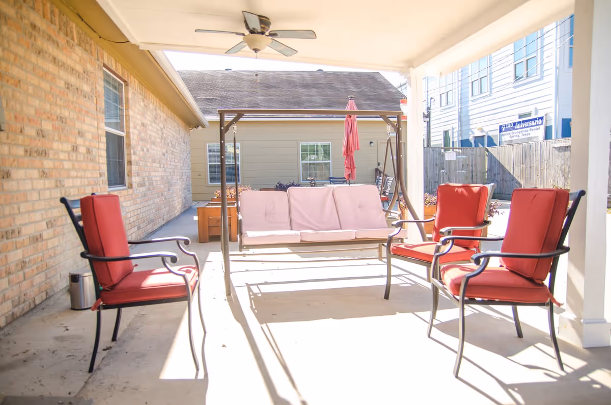 Covered outdoor patio area with a cushioned swing and four red cushioned chairs arranged around it. The patio is adjacent to a brick wall and overlooks a fenced yard with neighboring buildings visible in the background.