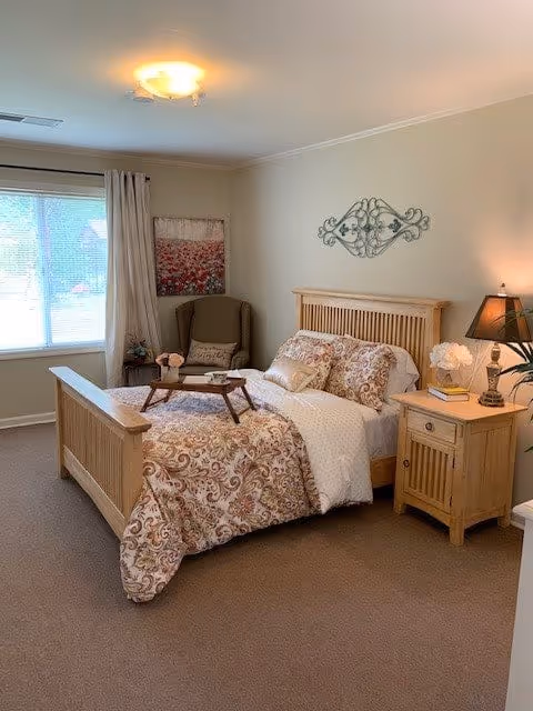 A cozy bedroom with a wooden bed frame and a patterned bedspread. There is a wooden nightstand with a lamp and decorative items next to the bed. A cushioned armchair is placed near a window with curtains, and a piece of wall art hangs above the chair. The room has beige walls and carpeted flooring.