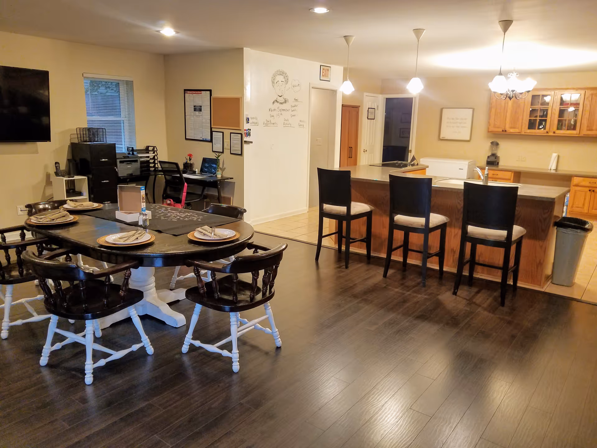Interior view of a senior living facility dining and kitchen area with a wooden dining table set for four, a kitchen island with three chairs, wooden cabinets, a wall-mounted TV, and a small office desk with a computer and printer in the background.
