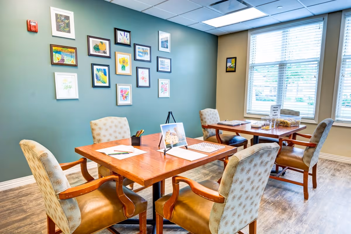A bright room with two wooden tables and six cushioned chairs arranged around them. The tables have papers, colored pencils, and a small display stand. The walls are decorated with multiple framed colorful artworks, and large windows with blinds let in natural light.