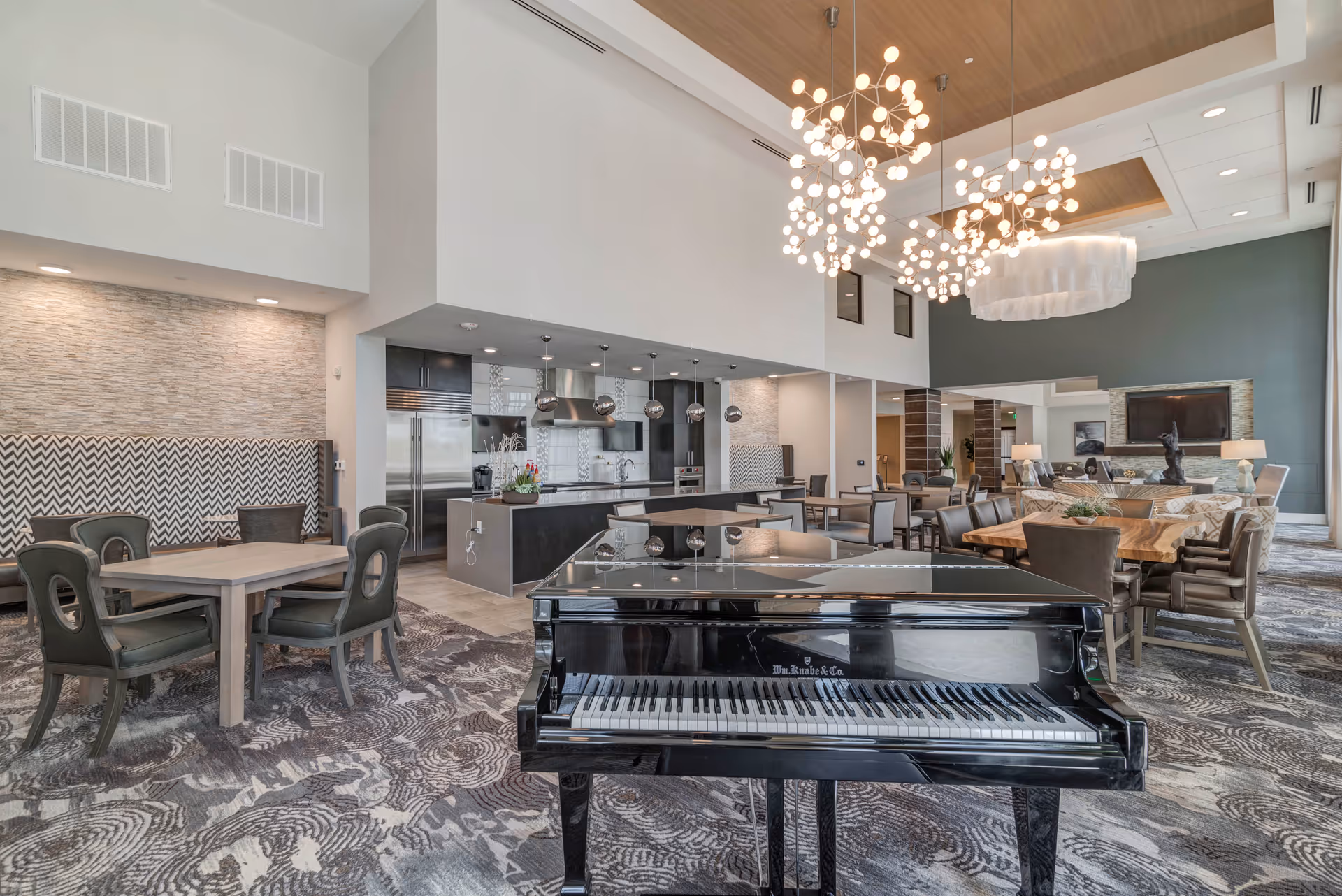 Spacious modern community lounge with a black grand piano in the foreground, dining tables, a kitchen area, and hanging chandeliers.