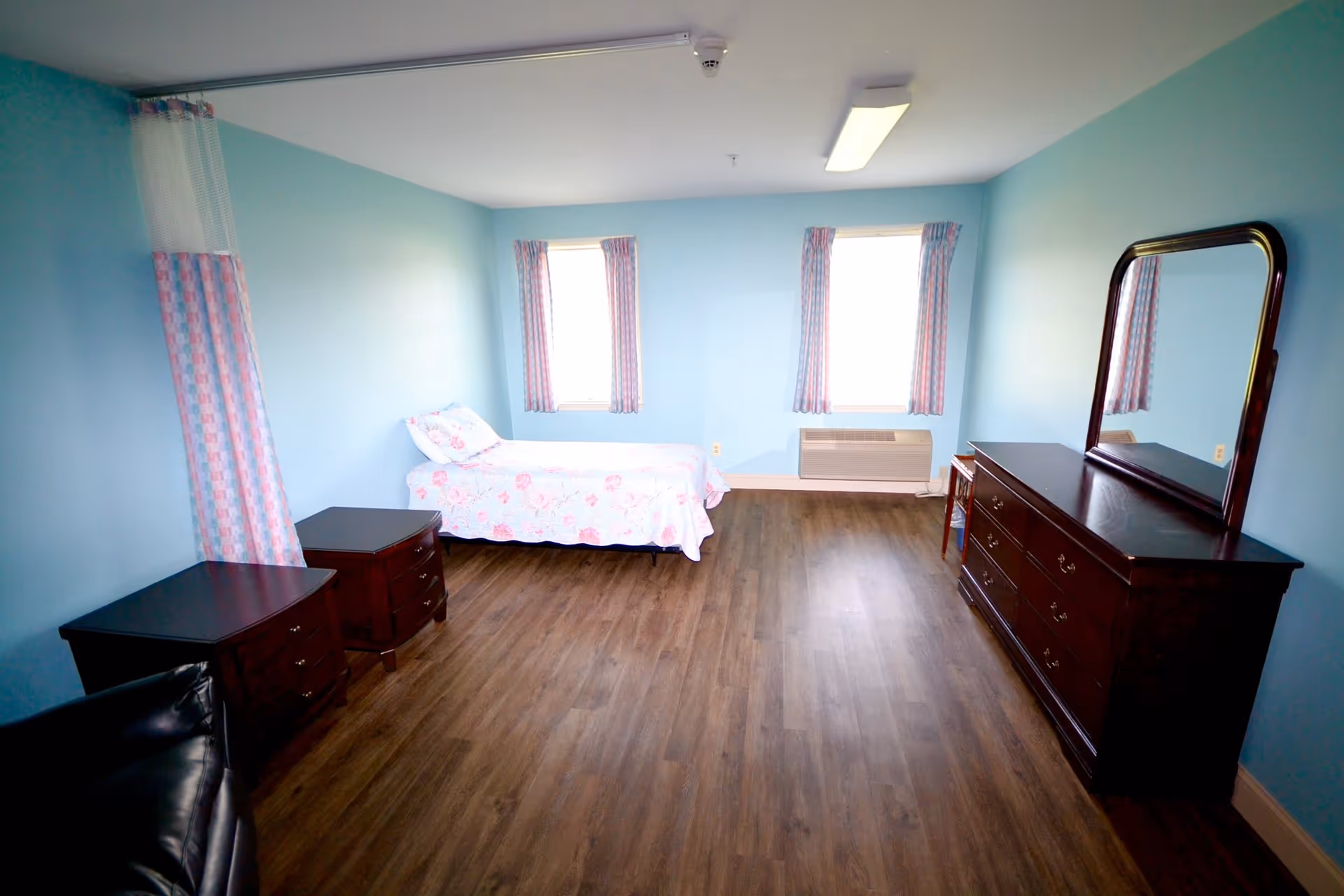 A simple bedroom with light blue walls and wood flooring. The room contains a single bed with floral bedding, two small dark wood nightstands, a large dark wood dresser with a mirror, a small chair, and two windows with patterned curtains. There is also a black leather chair partially visible in the foreground.