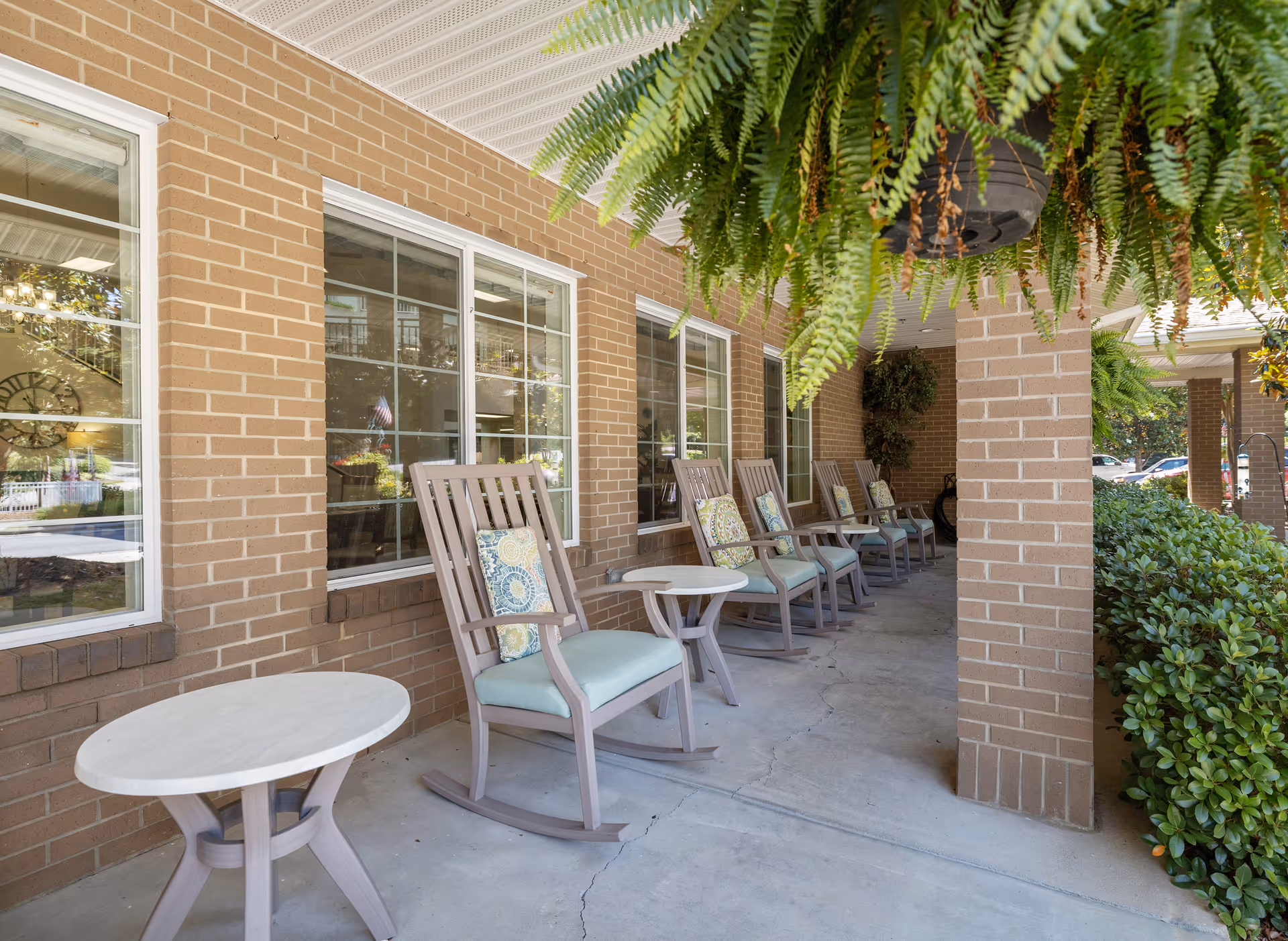 Covered outdoor patio area with several wooden rocking chairs each with a cushion and small round tables in front of them. The patio has brick walls with multiple windows and hanging green plants above. There are bushes and trees visible on the right side.