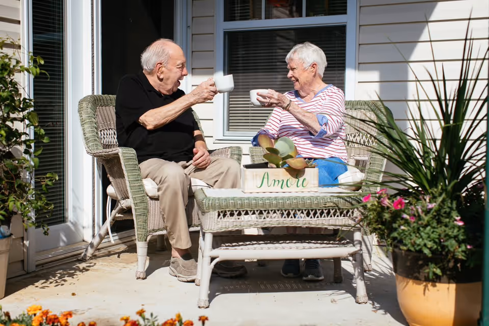 An elderly man and woman sitting on wicker chairs on a patio outside a building, smiling and clinking white coffee mugs together. There is a wicker table between them with a small plant and a wooden box labeled 'Amore'. The patio is decorated with potted plants and flowers.