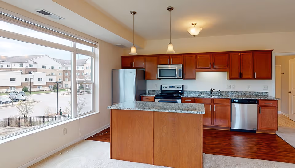A modern kitchen with wooden cabinets, granite countertops, and stainless steel appliances including a refrigerator, microwave, stove, and dishwasher. There is a kitchen island in the center with two pendant lights hanging above. Large windows on the left side provide natural light and a view of an apartment complex and parking area outside.