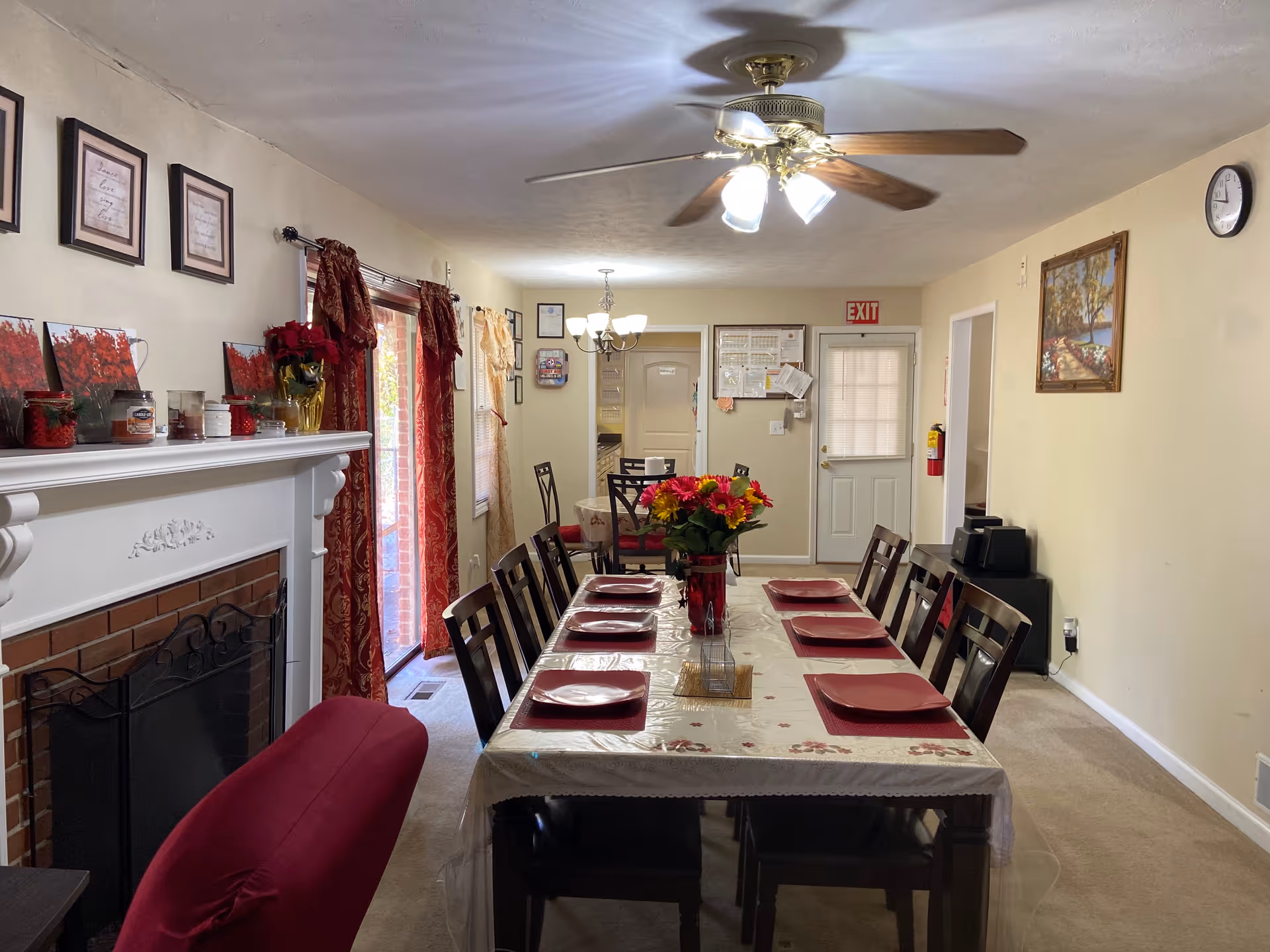 A dining room with a long table set with red plates and a floral centerpiece. The room features a ceiling fan with lights, a fireplace with decorative items on the mantel, red curtains on the windows, and framed pictures on the walls. There is a door at the far end with an exit sign above it.