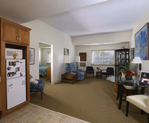 Spacious senior living suite interior with a small kitchenette and fridge on the left, a seating area with chairs and lamps, a dining table on the right, and a bedroom doorway in the back.