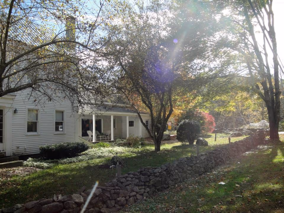 Exterior view of a white building with a covered porch and chairs, surrounded by trees and greenery with sunlight filtering through the leaves. A stone wall runs along the grassy area in front of the building.
