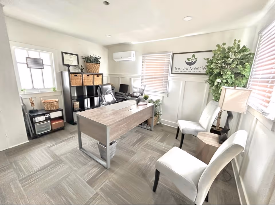 A bright office space with a wooden desk and black office chair. Behind the desk is a black shelving unit with wicker baskets and decorative items. Two white chairs and a small wooden side table with a lamp are positioned near the windows with blinds. A large potted plant is in the corner, and a framed sign on the wall reads 'Tender Mercies Compassionate Care'.