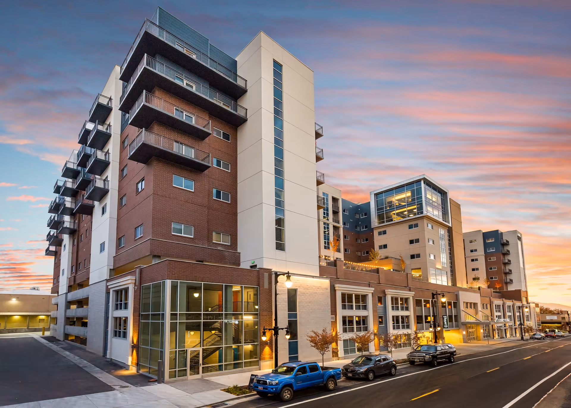 Exterior view of a multi-story senior living facility building at sunset with a partly cloudy sky. The building features a combination of brick and light-colored walls, multiple balconies, large windows, and street lamps. Several vehicles are parked along the street in front of the building.