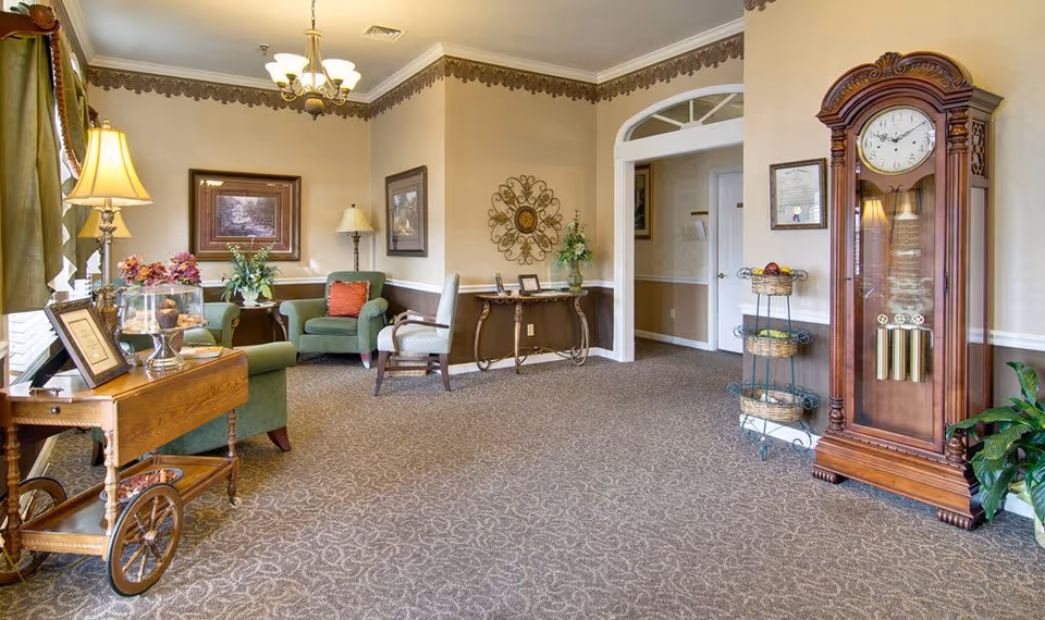 A cozy living room area with green upholstered chairs, a wooden side table with a lamp, framed artwork on the walls, a decorative wall piece, a wooden grandfather clock, and a small three-tiered fruit basket stand. The room has beige walls with a decorative border near the ceiling, carpeted floor, and a chandelier hanging from the ceiling.