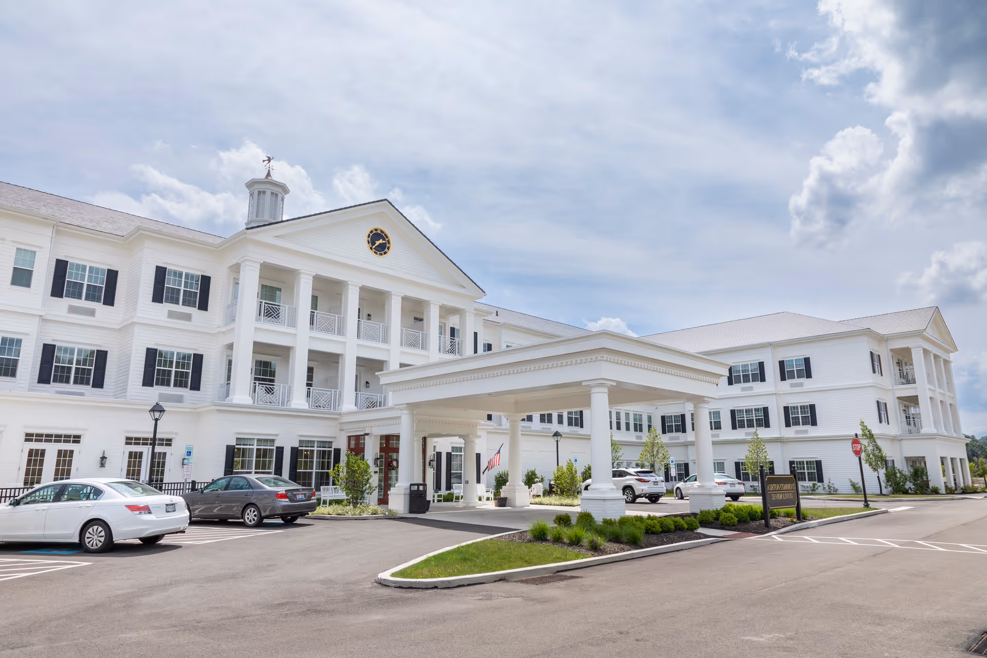 Exterior view of a large, white senior living facility building with multiple windows and black shutters. The building has a covered entrance supported by columns, a clock on the front gable, and a cupola on the roof. Several cars are parked in front of the building under a partly cloudy sky.