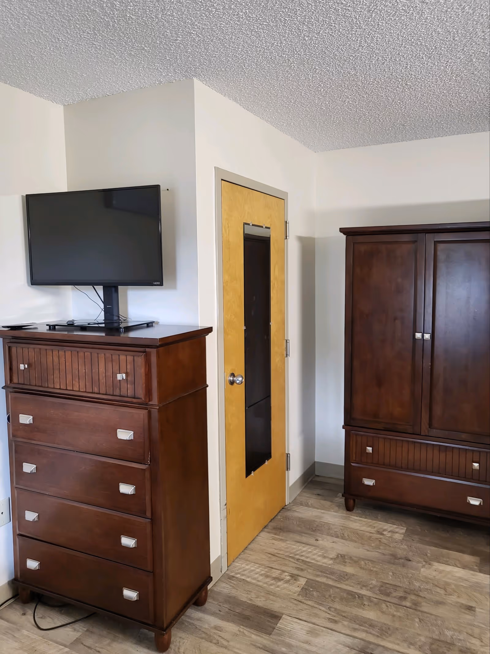 A corner of a room featuring a wooden dresser with a flat-screen TV mounted on the wall above it, a wooden armoire, and a wooden door with a vertical mirror attached. The floor is covered with wood-patterned vinyl or laminate flooring, and the walls are painted white.